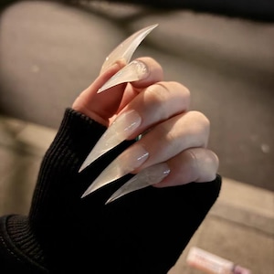 Op de afbeelding: Een hand met lange, heldere acrylnagels. De nagels zijn scherp gepunt, en steken ver voorbij de vingertoppen uit. De nagels hebben een glanzende afwerking, die licht reflecteert. De hand draagt een zwart shirt met lange mouwen.