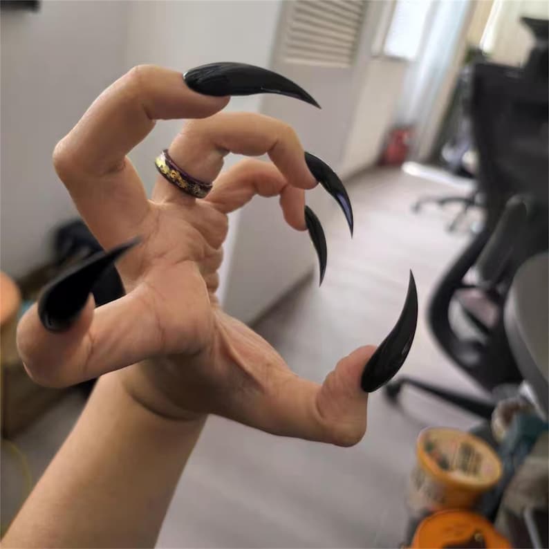 May include: A hand with long, glossy black fingernails, shaped like claws. A gold ring with a purple band is on the ring finger. The hand is in a curved position, with the fingers spread out.