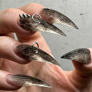 Op de afbeelding: Lange, puntige, heldere acrylnagels met een bruin, getextureerd patroon. Sommige nagels hebben zilveren ringen en puntige accenten, wat een unieke en edgy esthetiek creëert. De nagels zijn een statement piece.