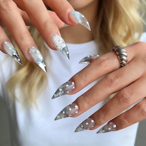 Puede incluir: Primer plano de uñas acrílicas largas y transparentes con forma puntiaguda. Las uñas presentan un diseño de purpurina plateada y lunares blancos. Se ve un anillo plateado en un dedo. Las uñas son un accesorio de moda.