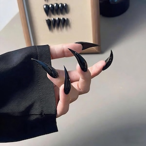 May include: A hand displaying long, glossy black stiletto nails. The nails are sharply pointed and curved, creating a dramatic and edgy appearance. The nails are a solid, opaque black colour. A black sleeve is visible in the background.