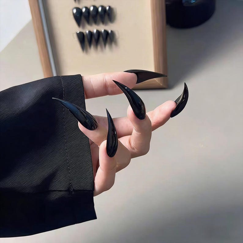 May include: A hand displaying long, glossy black stiletto nails. The nails are sharply pointed and curved, creating a dramatic and edgy appearance. The nails are a solid, opaque black colour. A black sleeve is visible in the background.