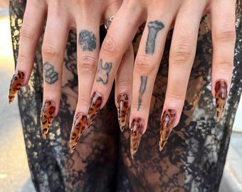 May include: Long, pointed fingernails with a leopard print design. The nails are a translucent amber colour with dark brown spots. The fingers have several small tattoos.