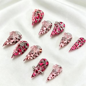 Peut inclure: Un ensemble de dix faux ongles avec un motif léopard rose et rouge. Certains ongles présentent des accents de paillettes argentées, des ornements floraux et de petits anneaux argentés. Les ongles sont présentés sur une surface blanche.