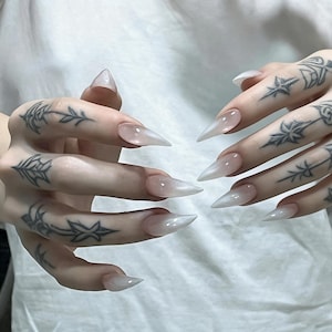Puede incluir: U&ntilde;as artificiales largas en forma de estilete con un acabado degradado blanco lechoso y transl&uacute;cido. Los dedos presentan intrincados tatuajes de tinta negra con dise&ntilde;os florales y de estrellas, mostrando una punta afilada en cada u&ntilde;a.