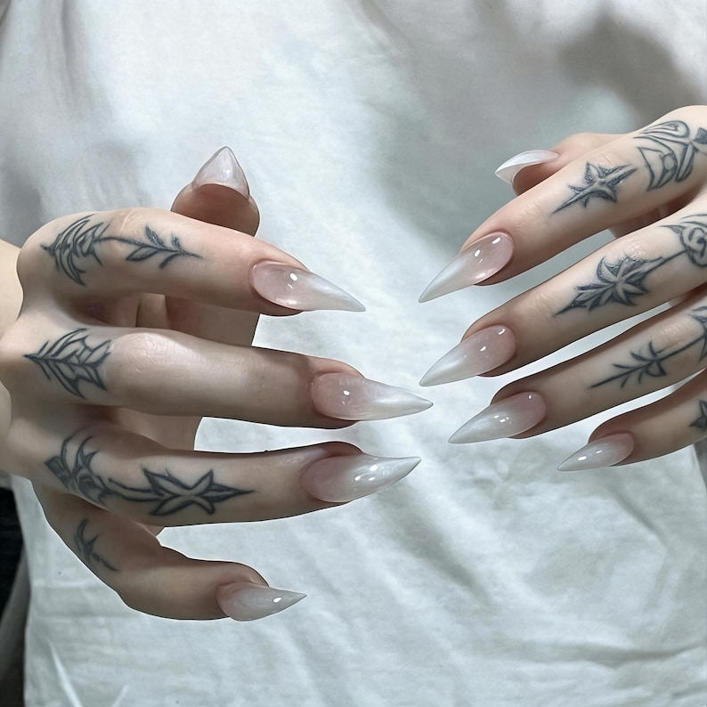 Puede incluir: U&ntilde;as artificiales largas en forma de estilete con un acabado degradado blanco lechoso y transl&uacute;cido. Los dedos presentan intrincados tatuajes de tinta negra con dise&ntilde;os florales y de estrellas, mostrando una punta afilada en cada u&ntilde;a.
