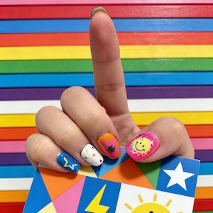 May include: A hand with colorful nail art, including a blue nail with a yellow star, a white nail with polka dots, and an orange nail with a green clover. The background is a rainbow of horizontal stripes.