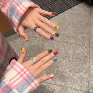 Pode incluir: Duas mãos com designs de unhas coloridos e artísticos. Cada unha apresenta uma combinação única de cores e formas, incluindo vermelho, verde, amarelo e azul. As mãos são adornadas com anéis, e as mangas têm um padrão xadrez rosa e branco.