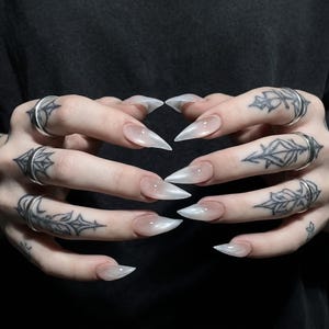 May include: Close-up of hands with long, pointed, pearlescent nails. The nails have a subtle gradient effect. The fingers are adorned with silver rings and intricate black tattoos. The background is a dark, solid colour.