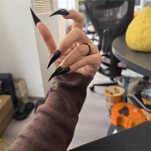 Può includere: Una mano con lunghe unghie nere a stiletto, un anello d'oro e una manica di maglione marrone. Le unghie sono appuntite e lucide. La mano fa il segno della pace.