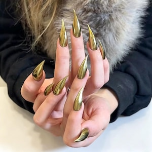 May include: A set of ten long, pointed, metallic gold press-on nails with a high-gloss, reflective finish. Each nail features a sharp, stiletto-like shape that tapers to a fine point, creating a smooth, uniform appearance across all fingers.