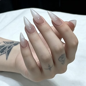 Puede incluir: Una mano con u&ntilde;as largas en forma de estilete puntiagudo que presentan un suave degradado blanco que se desvanece hacia una base natural transparente. La piel muestra peque&ntilde;os tatuajes de tinta negra en la mu&ntilde;eca y los dedos, con un acabado pulcro.