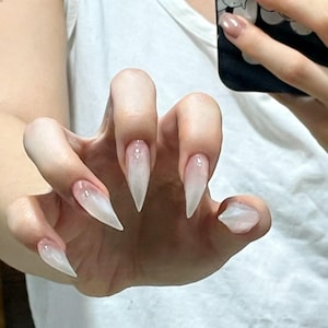 Puede incluir: U&ntilde;as largas en forma de estilete puntiagudo con un acabado blanco lechoso transl&uacute;cido. Las u&ntilde;as presentan una textura suave y brillante que capta la luz, creando un sutil efecto degradado desde la base hasta las puntas afiladas.