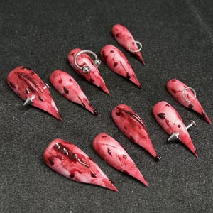 May include: A set of ten long, almond-shaped, artificial nails. The nails are painted with a pinkish-white base and splattered with dark red, resembling blood. Some nails feature silver-colored metal accents, including rings and studs.