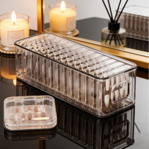 Può includere: Organizzatore per unghie in plastica trasparente di forma rettangolare con coperchio. L'organizzatore è pieno di unghie finte. Un organizzatore più piccolo e quadrato con unghie è di fronte. Candele accese e un diffusore sullo sfondo.