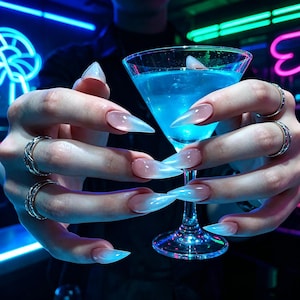 May include: A martini glass filled with a vibrant blue liquid is held by hands adorned with long, pointed, ombre-colored nails. Silver rings decorate the fingers. The background features neon lights.