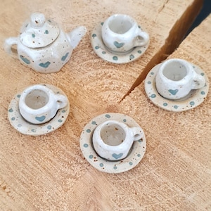 Könnte beinhalten: Ein Miniatur-Teeservice aus Keramik mit einer Teekanne und drei Tassen mit Untertassen. Das Set ist cremefarben mit blauen Herz- und Punktakzenten. Das Teeservice wird auf einer hellbraunen, strukturierten Oberfläche präsentiert.