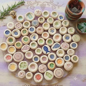 Könnte beinhalten: Eine Sammlung kleiner, runder, cremefarbener Keramikknöpfe mit verschiedenen geprägten Motiven wie Blumen, Muscheln, Fischen und geometrischen Mustern. Die Knöpfe sind auf einer transparenten Oberfläche angeordnet, mit einem Bienenstempel.