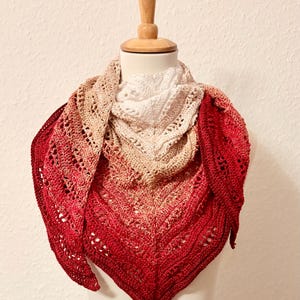 Puede incluir: Un chal de ganchillo hecho a mano en forma triangular, exhibido en un maniquí. El chal presenta un degradado de colores, que va del blanco al beige y finalmente al rojo intenso. El diseño calado añade un toque delicado.