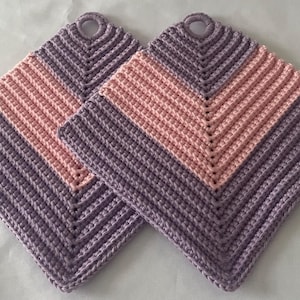 May include: Two square crochet potholders featuring a chevron pattern in alternating bands of light purple and soft pink. Each potholder is constructed with a textured stitch and includes a circular loop at the top corner for convenient hanging.