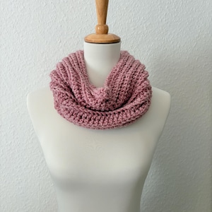 Op de afbeelding: Een stoffig roze, dik gebreide infinity sjaal, tentoongesteld op een witte mannequin. De sjaal is gemaakt van dik garen met een geribbelde textuur. De mannequin staat tegen een witte, getextureerde muur.