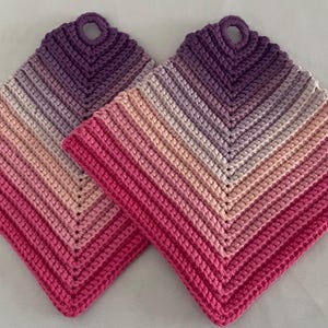 Può includere: Due presine quadrate fatte a mano all'uncinetto. Le presine presentano una sfumatura di colori, che passa dal rosa intenso al rosa chiaro, poi al lavanda e infine al viola scuro nella parte superiore. Ogni presina ha un anello per appenderla.