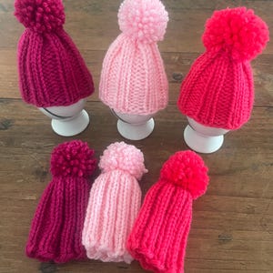 Könnte beinhalten: Sechs gestrickte Eierwärmer in den Farben Magenta, Hellrosa und leuchtendem Rot. Jeder Eierwärmer hat eine gerippte Struktur und einen passenden Wollbommel an der Spitze. Drei Eierwärmer stehen auf weißen Eierbechern, drei liegen flach.