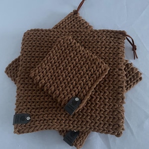 Può includere: Un set di tre articoli marroni all'uncinetto, tra cui due sacchetti quadrati e un articolo rettangolare più piccolo. I sacchetti hanno una trama strutturata ed etichette in pelle. Un sacchetto ha un cordoncino in pelle. Gli articoli sono disposti su una superficie bianca.
