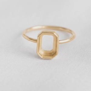 May include: A gold ring featuring a thin, polished band and a central, empty rectangular setting with beveled edges. The setting is designed to hold a stone, but it is currently vacant, showcasing the smooth, metallic finish of the gold structure.