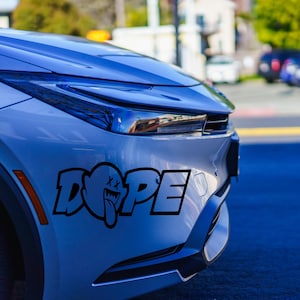 May include: A close-up of a light blue car's front side, featuring a black sticker that reads "DOPE" in a stylized font. The sticker includes a ghost-like character with crossed-out eyes and a protruding tongue. The car's design is modern.