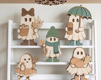 May include: Five layered wooden ghost cutouts displayed on a white shelf. The ghosts feature autumn-themed accessories including a basket of apples, a wheat bundle, a green beanie and scarf with a lantern, a coffee cup and umbrella, and pumpkins.