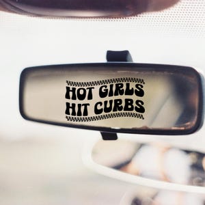 May include: A car's rearview mirror with the text "HOT GIRLS HIT CURBS" in bold, black letters. The text is framed by a tire track design. The mirror is rectangular and mounted on a black stem.