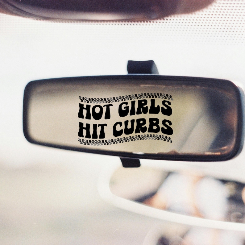 May include: A car's rearview mirror with the text "HOT GIRLS HIT CURBS" in bold, black letters. The text is framed by a tire track design. The mirror is rectangular and mounted on a black stem.