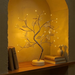Könnte beinhalten: Eine dekorative baumförmige Lampe mit goldfarbenen Blättern und kleinen, warmweißen LED-Leuchten. Die Lampe steht auf einem weißen Sockel und wird auf einem Holzregal mit Büchern und einer kleinen Vase ausgestellt. Der Hintergrund ist eine warmgelb getönte Wand.