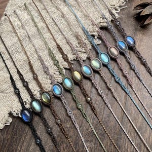 May include: A collection of macrame bracelets with oval labradorite stones. The bracelets feature various cord colors, including black, brown, green, and blue. The labradorite stones have a shimmering, iridescent quality, and the bracelets are displayed on a textured surface.