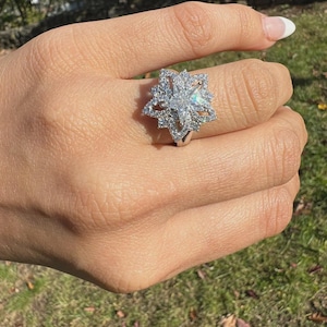 May include: A silver ring with a large, starburst design. The ring features a central, clear gemstone surrounded by smaller, sparkling stones. The ring is worn on a hand with a manicured nail.
