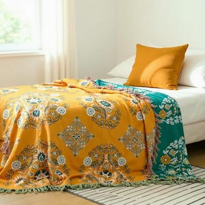 Puede incluir: Una manta de color amarillo mostaza con un patrón floral detallado y un borde turquesa está sobre una cama. Una almohada naranja descansa sobre la cama. La manta tiene un ribete de flecos.