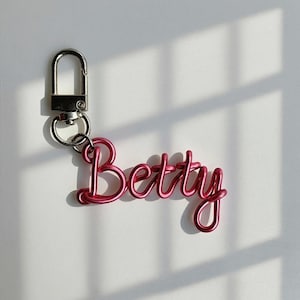 Può includere: Un portachiavi in filo rosa con la scritta "Betty" in un carattere corsivo. Il portachiavi ha un fermaglio argentato e un piccolo anello. Lo sfondo è una superficie bianca liscia con ombre.