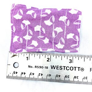 May include: A small, rectangular lavender fabric item with a white floral pattern. The item is approximately 3 inches long, as measured by a ruler. The fabric has a soft, slightly wrinkled appearance.