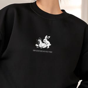 Può includere: Felpa nera con una grafica bianca di una gallina e un coniglio, con la scritta "HOW EASTER EGGS ARE REALLY MADE" sotto. La felpa ha un girocollo e maniche lunghe.