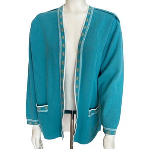 May include: A teal blue cardigan with white and gold trim. The cardigan has a V-neck and long sleeves. It has two pockets on the front.