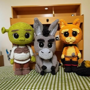 May include: Three handmade crochet dolls featuring a green ogre with a white tunic, a grey donkey with black accents, and an orange cat with a black cape and boots. Each figure has large black plastic eyes and detailed yarn stitching throughout.
