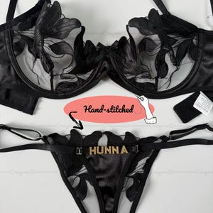 Puede incluir: Conjunto de lencería negra con sujetador y tanga. El sujetador tiene detalles de encaje floral y aros. El tanga tiene la palabra "HUNNA" en letras doradas. El conjunto está cosido a mano.