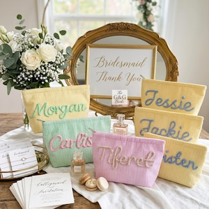May include: A collection of pastel-colored cosmetic bags with embroidered names, including "Morgan," "Carlis," "Tiferet," "Jessie," "Jackie," and "Kristen." The bags are arranged with a gold-framed mirror, a bouquet of white flowers, and a sign that reads "Bridesmaid Thank You."