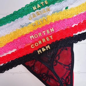 Könnte beinhalten: Verschiedene String-Tangas in Grün, Weiß, Gelb, Pink, Rot und Schwarz. Jeder String hat eine Spitzenborte und goldene Buchstaben, die Namen oder Initialen buchstabieren. Die Unterwäsche ist aus zartem, durchsichtigem Stoff gefertigt.