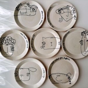 May include: Seven round, off-white ceramic plates arranged in a cluster. Each plate features a unique, simple black line drawing of a face or animal, accompanied by handwritten black text phrases such as 'there are others' and 'not rated for buffets'.
