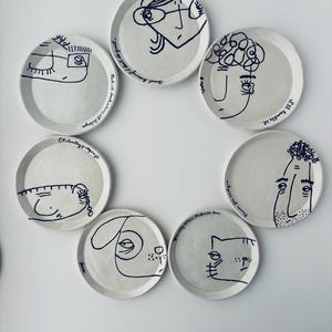 Op de afbeelding: Een set van zeven witte keramische borden met zwarte lijntekeningen van gezichten en dieren. Elk bord heeft een uniek ontwerp, met tekst zoals "I'll handle it" en "Put it back where it belongs."