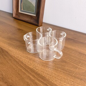 Puede incluir: Cuatro pequeñas tazas de vidrio transparente con asas están dispuestas sobre una superficie de madera. Las tazas tienen un diseño simple y minimalista. Una imagen enmarcada con un paisaje está en el fondo.