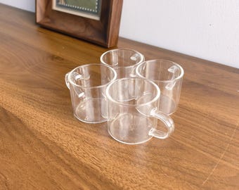 May include: Four small, clear glass cups with handles are arranged on a wooden surface. The cups have a simple, minimalist design. A framed picture with a landscape scene is in the background.