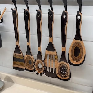 May include: A set of six wooden kitchen utensils hanging from a black metal rack. The utensils feature a distinct layered wood grain pattern with alternating dark brown and light tan bands, including a spatula, spoon, pasta server, and slotted turner.